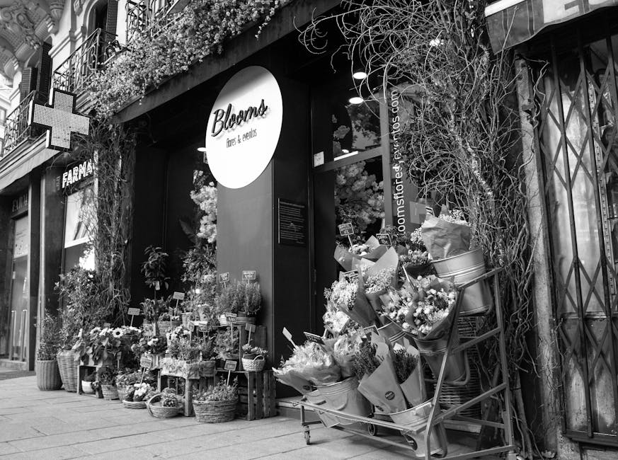 A black-and-white image of the storefront for Bloom's flowers & gifts, featuring an outdoor display of fresh flowers arranged in baskets, crates, and buckets. The floral arrangements include various t