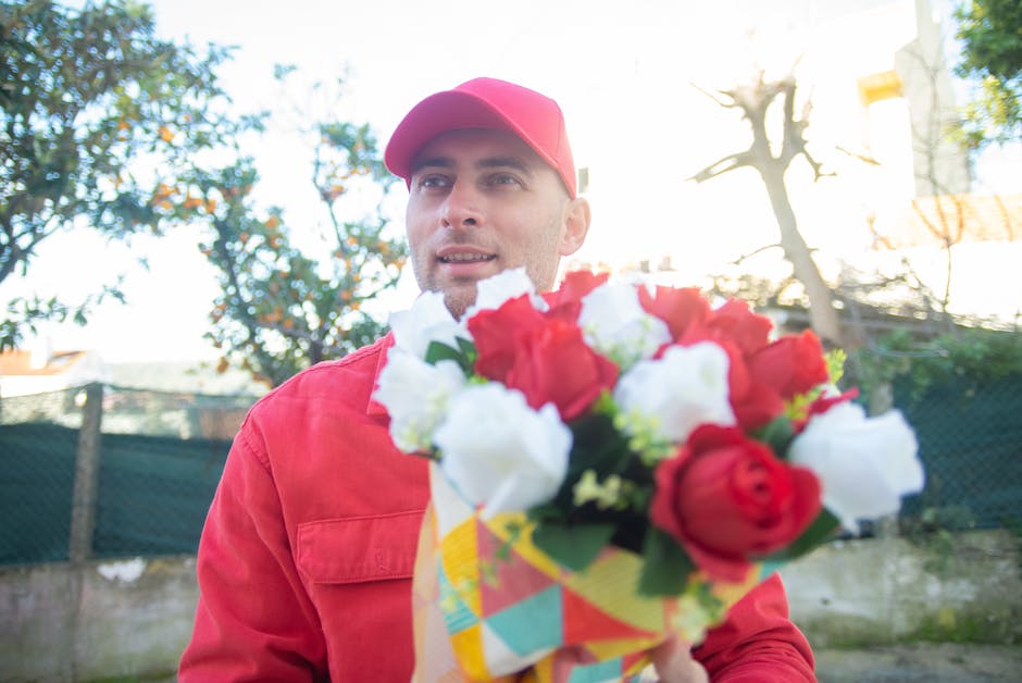 A male flower delivery courier from flowerswinchmorehill.co.uk, dressed in a red jacket and matching red cap, is holding a vibrant bouquet of fresh flowers close to the camera. The bouquet features a 