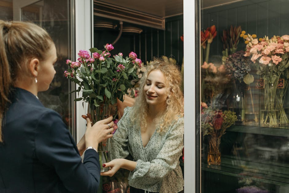A woman with curly blonde hair, dressed in a light-colored floral blouse, smiling gently as she receives a tall bouquet of fresh pink roses from a florist wearing a dark blazer. The florist's hands ar