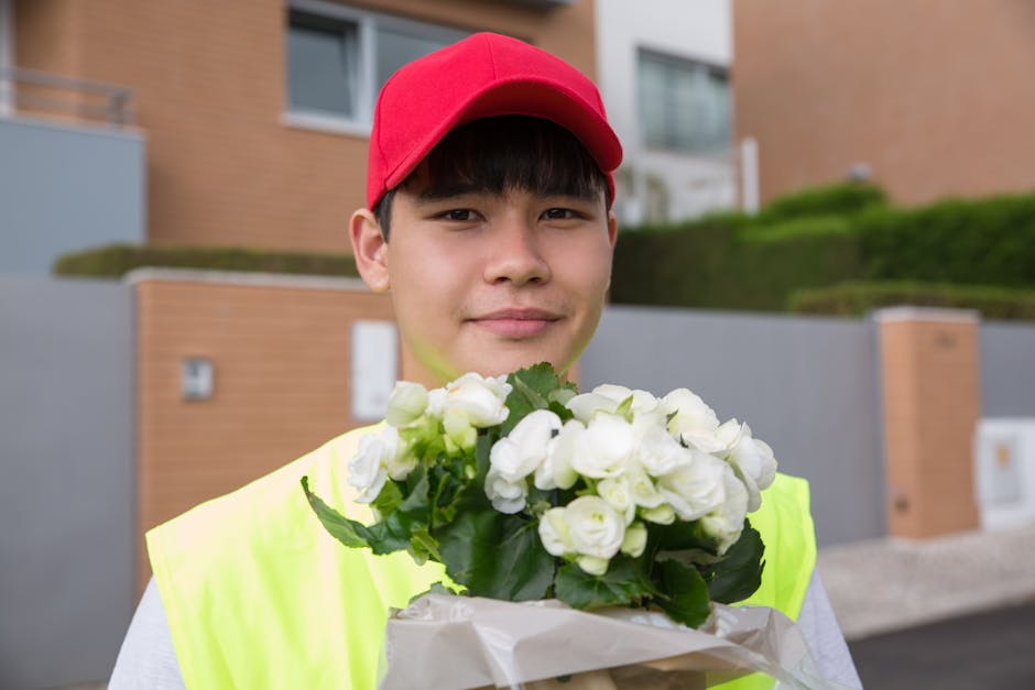 A young male flower delivery courier wearing a red cap and a high-visibility yellow vest is holding a bouquet of fresh white roses wrapped in white paper. His expression is friendly and welcoming, wit
