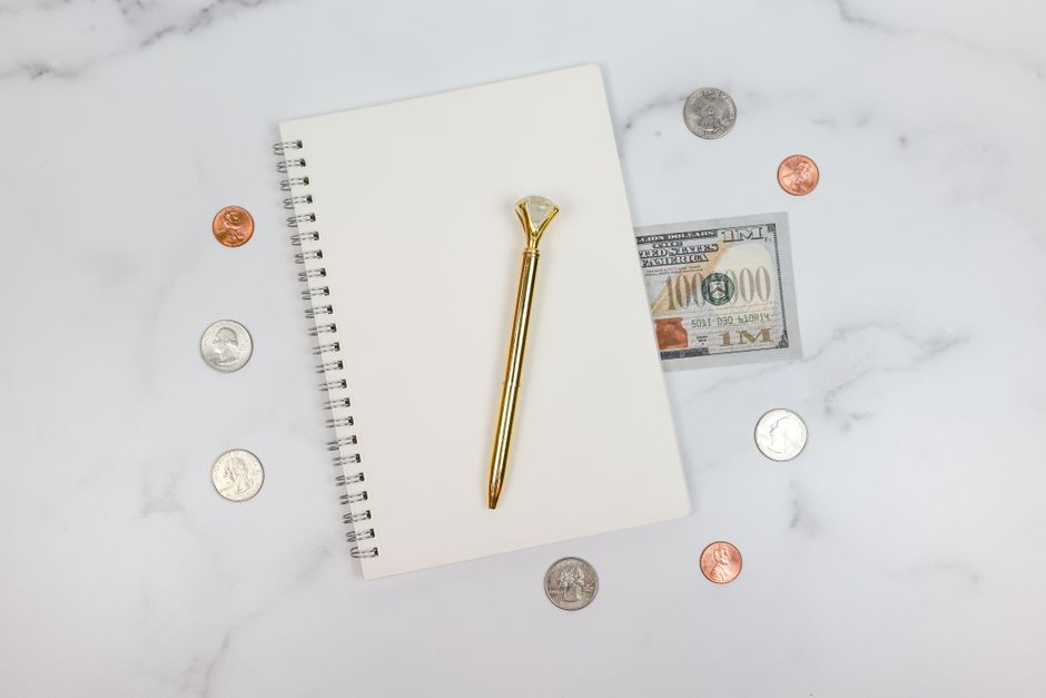 A white spiral-bound notepad with blank pages is lying flat on a marble surface, accompanied by a gold-colored ballpoint pen placed diagonally across the notebook. Surrounding the notepad are various 