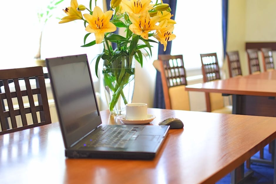 A clear glass vase filled with vibrant yellow lilies and green foliage, arranged in a bouquet on a polished wooden table. The bouquet features large, trumpet-shaped blossoms with orange and dark strea