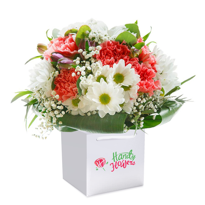Expertly crafted and elegantly upright in a clean white square gift bag, this bouquet brings together the serene beauty and festivity cherished in Winchmore Hill's floral traditions. A full, domed mix of coral, peach, and blush red carnations serves as the centrepiece, each flower's frilled petals offering a dense, almost cloudlike softness. Bright, white daisy-form blooms with vivid green centres dance among the carnations, lending a youthful charm reminiscent of springtime walks along Church Hill. Soft clusters of white gypsophila thread lightly throughout, diffusing light and adding layers of feathery texture. Tucked within the mix, a few deep-hued purple buds stand unopened, their presence adding depth, dimension, and anticipation of coming blooms. Surrounding the florals, an abundance of glossy, deep green leaves frames the bouquet, snugly cradling the arrangement in a natural embrace. Satin ribbon handles crown the sturdy gift bag, promising ease of delivery or presentation for a birthday, anniversary, or meaningful moment. Set against a flawless white backdrop, every nuance of colour, texture, and florist craftsmanship is clearly visible-making this image an ideal representation of Winchmore Hill's flower delivery artistry and attentive service. The careful balance of vibrancy, elegance, and freshness echoes the atmosphere of the N21 community.